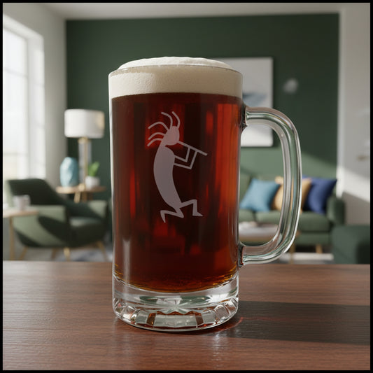 A clear glass beer mug with a handle, filled with dark amber beer and topped with white foam, featuring a etched kokopelli figure. The mug is placed on a light wood surface with a living room in the background.