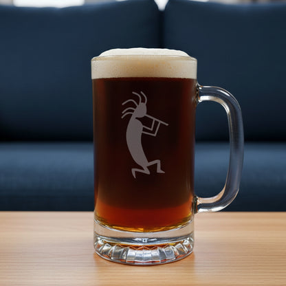 A clear glass beer mug with a handle, filled with dark amber beer and topped with white foam, featuring a etched kokopelli figure. The mug is placed on a light wood surface with a blue couch in the background.
