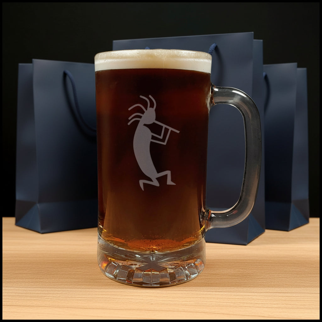 A clear glass beer mug with a handle, filled with dark amber beer and topped with white foam, featuring a etched kokopelli figure. The mug is placed on a light wood surface with a gift bags in the background.