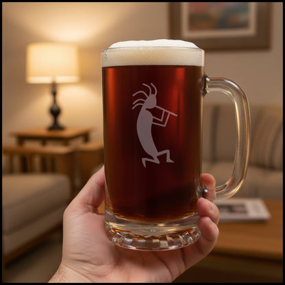 A clear glass beer mug with a handle, filled with dark amber beer and topped with white foam, featuring a etched kokopelli figure. The mug is placed on a light wood surface with a living room in the background.