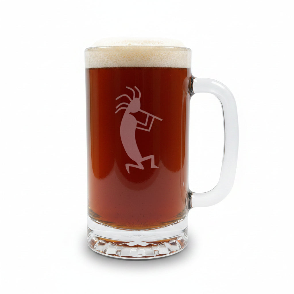 A clear glass beer mug with a handle, filled with dark amber beer and topped with white foam, featuring a etched kokopelli figure. The mug is placed on a light wood surface with a white background.
