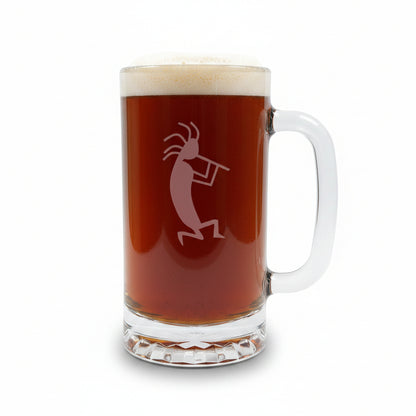 A clear glass beer mug with a handle, filled with dark amber beer and topped with white foam, featuring a etched kokopelli figure. The mug is placed on a light wood surface with a white background.