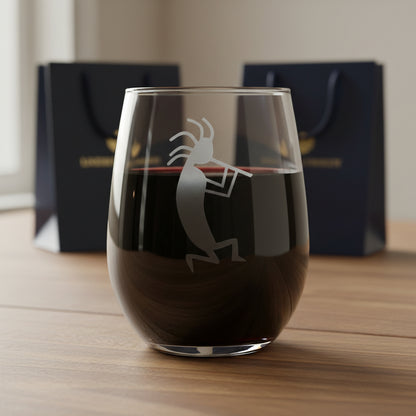 A clear stemless wine glass with an etched Kokopelli dancer design, resting on a wooden surface with 2 gift bags in the  background.