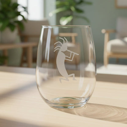 A clear stemless wine glass with a white etched Kokopelli dancer design, sitting on a light wooden table in a softly blurred living room setting.