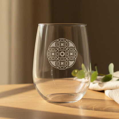 A clear stemless wine glass with a etched mandala design on the front, set on a wooden table in warm natural sunlight with blurred green leaves and a neutral background.