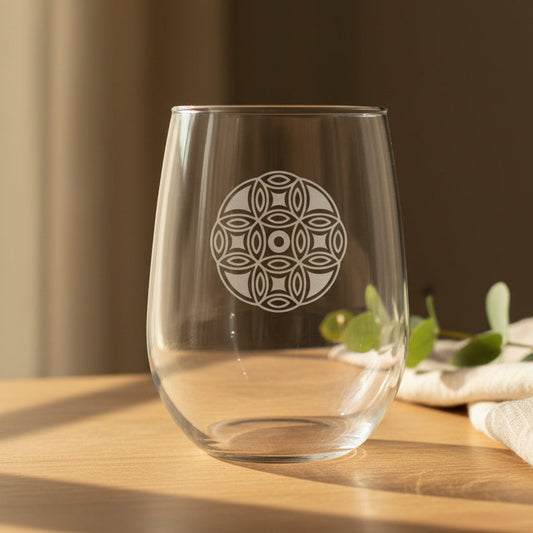 A clear stemless wine glass with a etched mandala design on the front, set on a wooden table in warm natural sunlight with blurred green leaves and a neutral background.