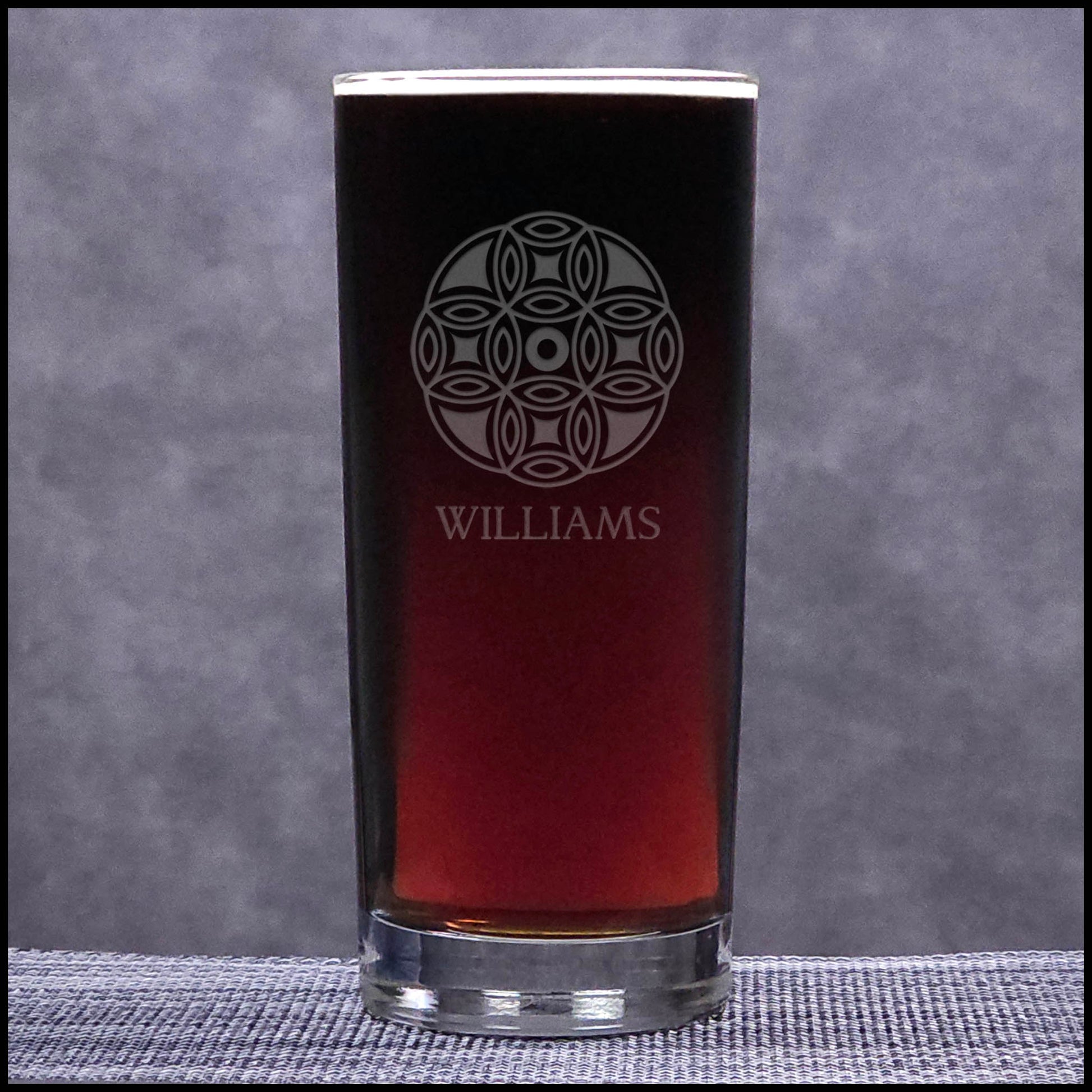 A clear cooler glass filled with dark red drink , featuring an etched circular mandala pattern and the name 'WILLIAMS' etched on the front, set against a textured gray background.