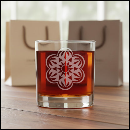 A clear glass rocks tumbler filled with amber liquor, featuring a white etched floral mandala design on the front, set on a wooden table with blurred beige gift bags in the background.