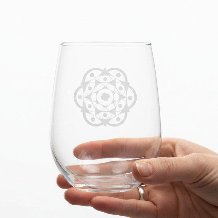 A clear stemless wine glass with a frosted geometric mandala design, held by a person's hand against a plain white background.