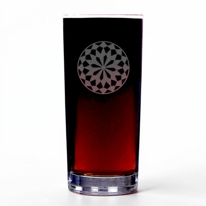 A clear highball glass with a red brown beverage, featuring a circular geometric mandala pattern on the upper half, set against a plain white background.