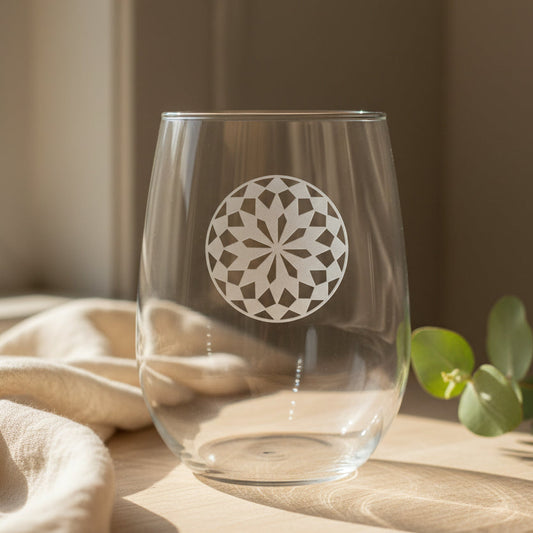 A clear stemless wine glass with a mandala design sand etched on the front, resting on a wooden surface against a neutral background.