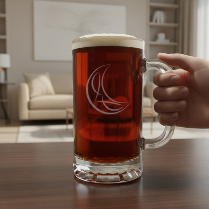 A large clear glass beer mug filled with amber beer and topped with white foam, held by a hand on a wooden table in a blurred living room setting. The mug has a white etched crescent design.