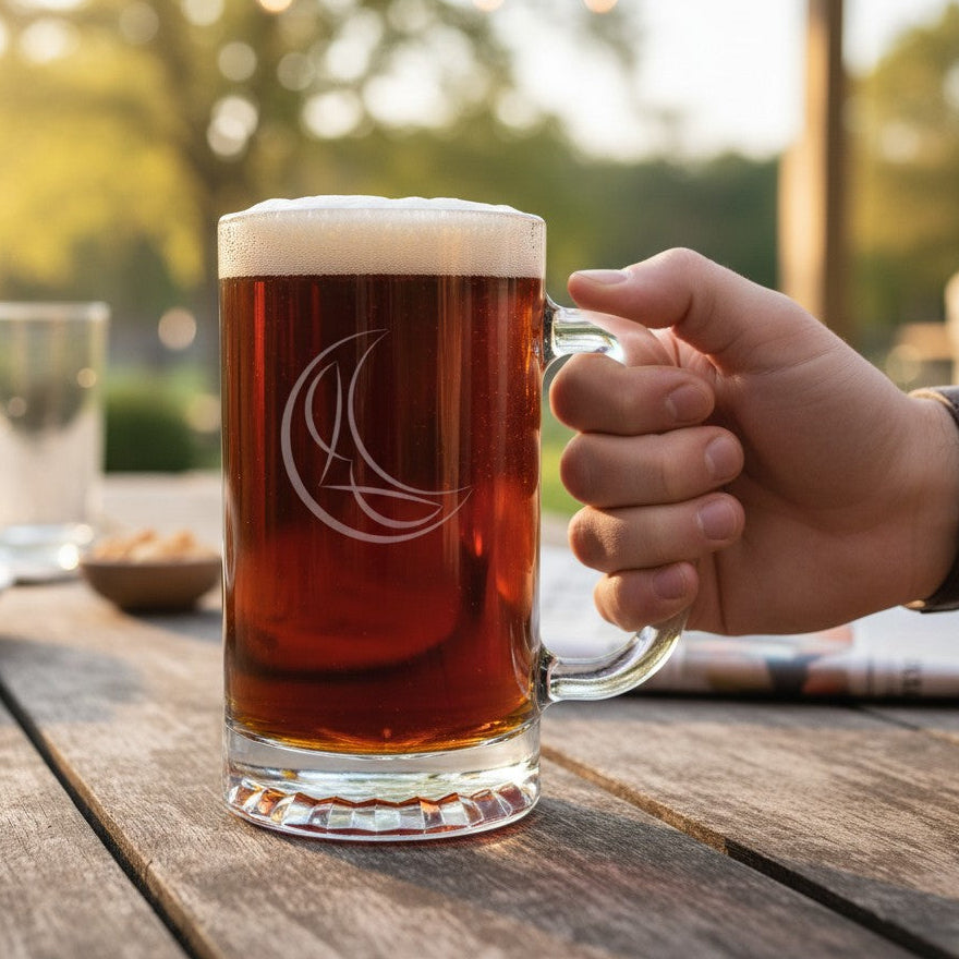 A hand holds an etched glass beer mug filled with amber beer and topped with white foam, set on a wooden table outdoors with a blurred background.