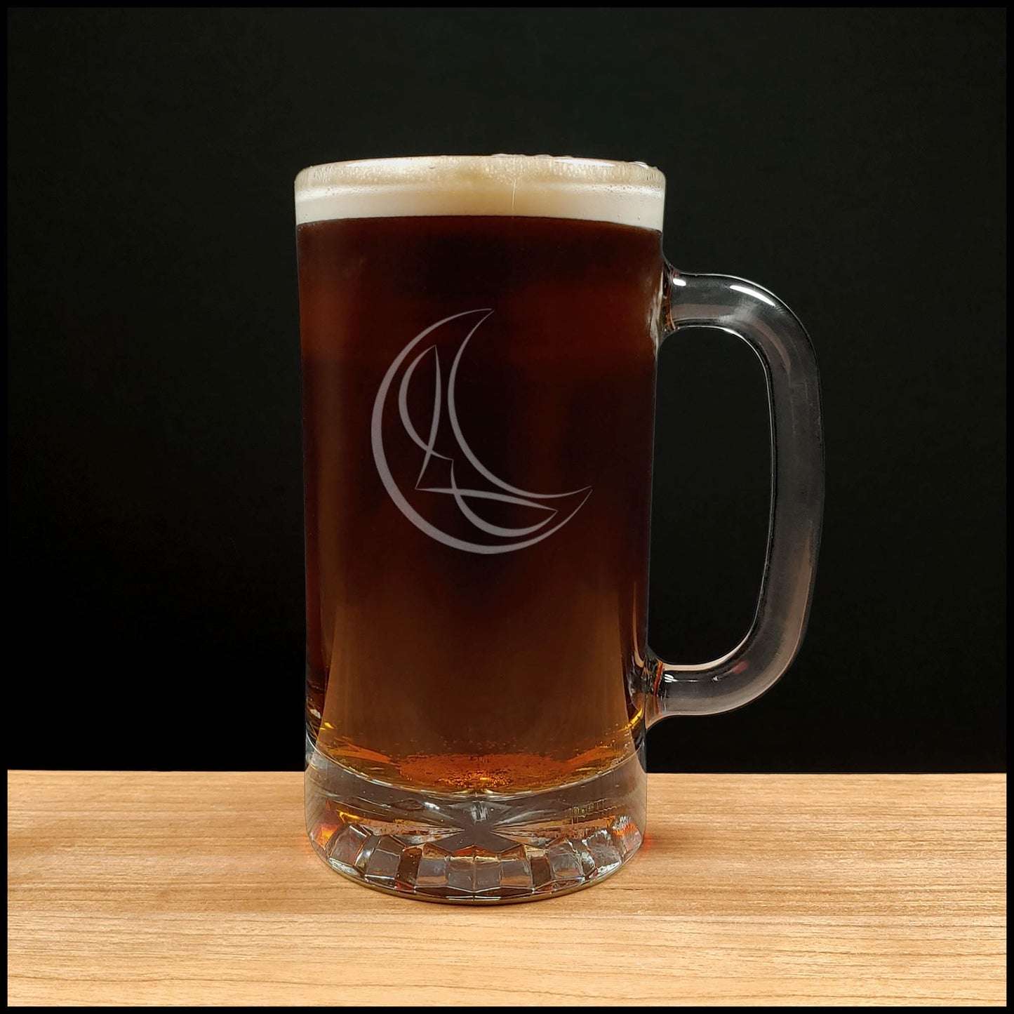 A clear glass beer mug with a handle, filled with dark amber beer and topped with white foam, standing on a light wood surface against a black background. The mug has an etched crescent moon and stylized letter design.