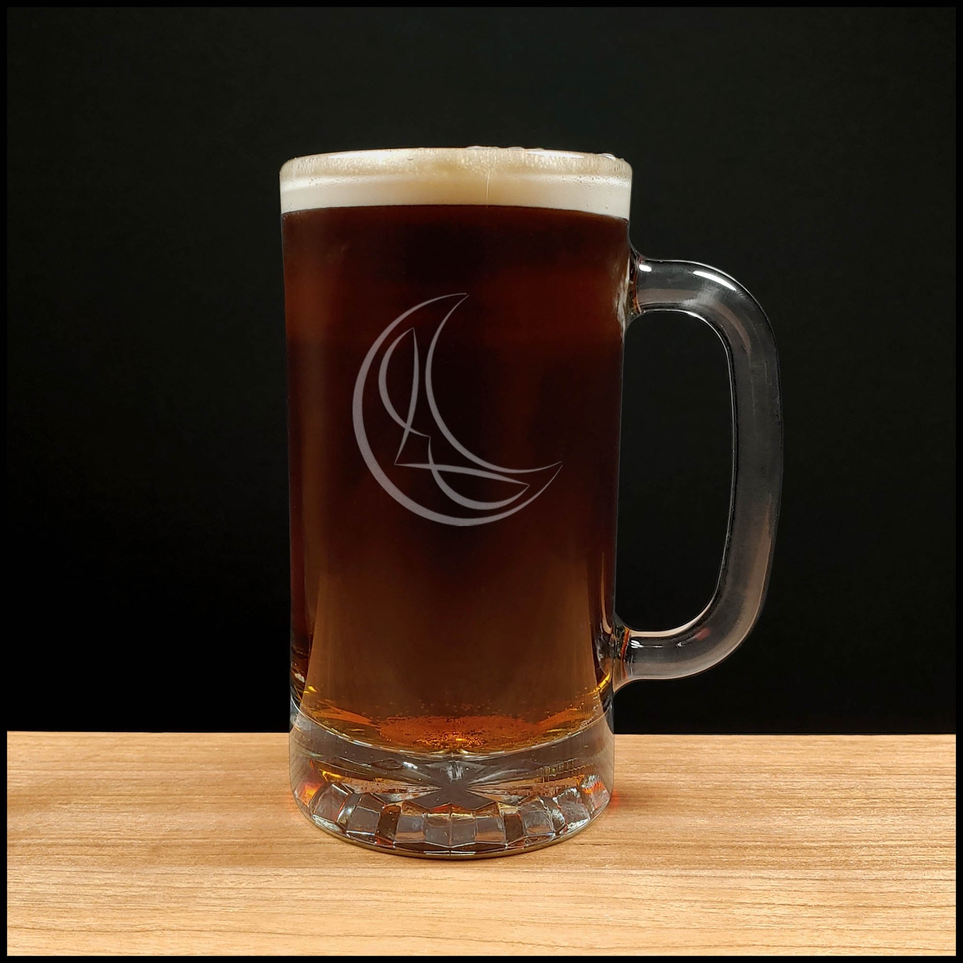 A clear glass beer mug with a handle, filled with dark amber beer and topped with white foam, standing on a light wood surface against a black background. The mug has an etched crescent moon and stylized letter design.
