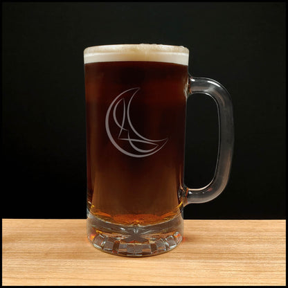 A clear glass beer mug with a handle, filled with dark amber beer and topped with white foam, standing on a light wood surface against a black background. The mug has an etched crescent moon and stylized letter design.