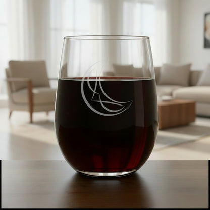 A clear stemless wine glass with a white crescent moon and abstract line design etched on the front, resting on a wooden surface 