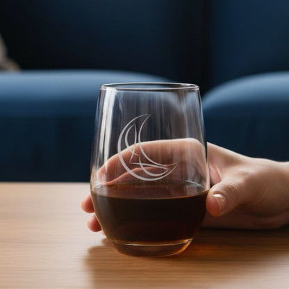 A hand holds a clear stemless wine glass with an etched crescent moon design, filled with a dark red beverage, set on a wooden table with a blurred blue sofa in the background.