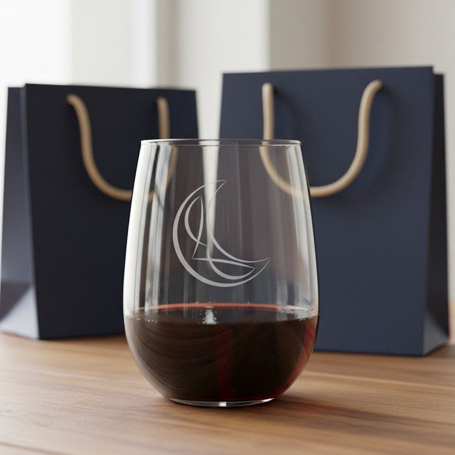 A clear stemless wine glass with an etched crescent moon design, partially filled with red wine, set on a wooden table with two navy gift bags in the background.