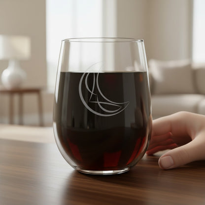 A clear stemless wine glass with an etched crescent moon and star design, partially filled with dark red wine, sitting on a wooden table with a hand resting near the rim in a softly blurred living room setting.
