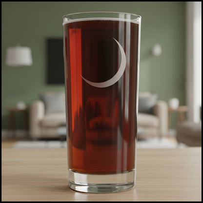 A tall, clear pint glass filled with a very dark reddish-brown liquid, featuring a etched crescent moon design on the front, set against a textured gray background.