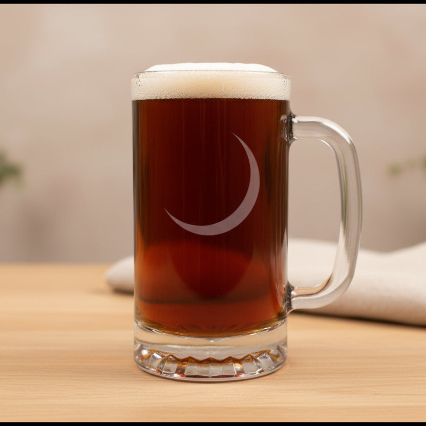 A clear glass beer mug with a handle, filled with amber beer and topped with white foam, featuring an etched crescent moon on the front, set on a wooden table with a soft beige background.