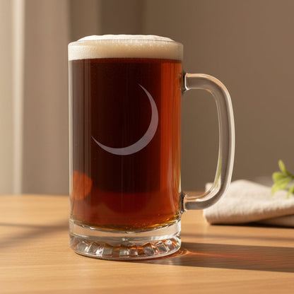 A clear glass beer mug with a handle, filled with dark beer and topped with white foam, featuring an etched crescent moon design on the front, set on a wooden surface against a black background.