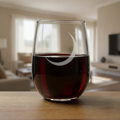 A clear stemless wine glass with a gray crescent moon design on the front, resting on a wooden surface against a cozy living room background.