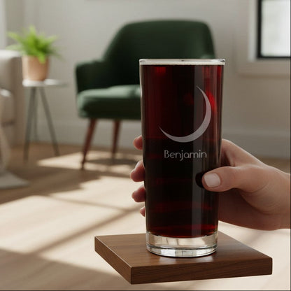 A hand holds a clear glass tumbler with a dark red drink and a white crescent moon and 'Benjamin' logo, resting on a square wooden coaster in a softly blurred living room setting.