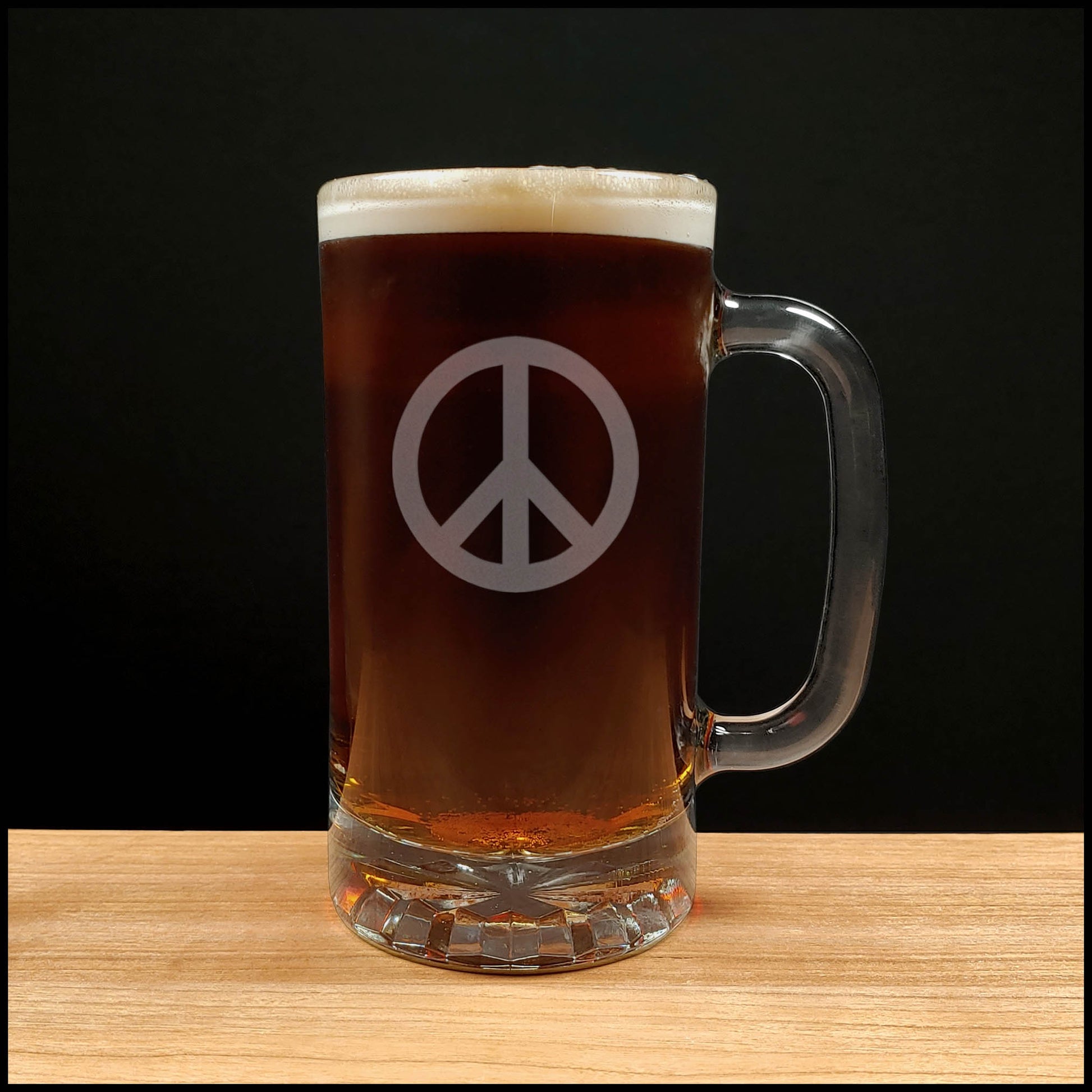 A clear glass beer mug filled with dark beer, featuring a frosted white peace sign on the front, set on a light wood surface against a black background.