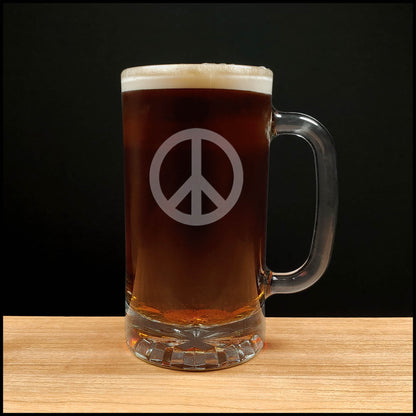 A clear glass beer mug filled with dark beer, featuring a frosted white peace sign on the front, set on a light wood surface against a black background.