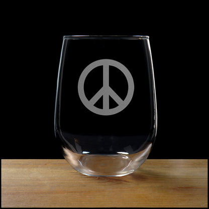 A clear stemless wine glass with a gray peace sign etched on the front, resting on a wooden surface against a solid black background.