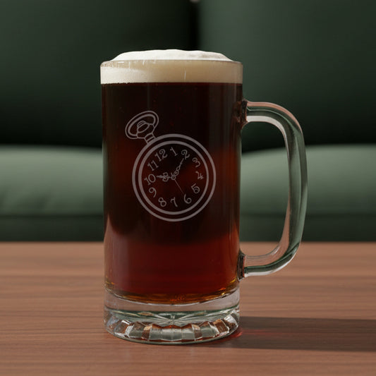 A clear glass beer mug with a handle, filled with dark amber beer and topped with white foam, featuring an etched pocket watch design on the front, set on a light wood surface against a green couch background.