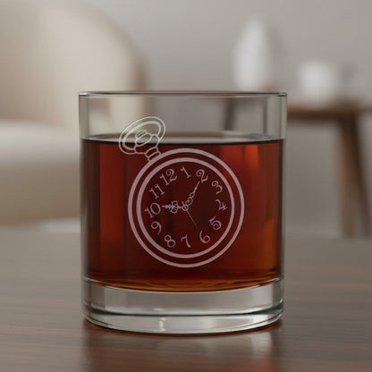 A clear lowball glass filled with amber-brown liquid, featuring an etched pocket watch on the front, set on a wooden table with a softly blurred indoor background.