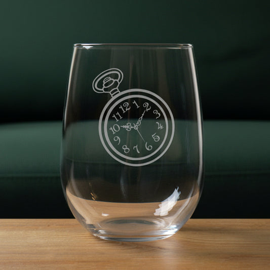 A clear stemless wine glass with an etched pocket watch design on the front, resting on a wooden surface against a green couch in the background