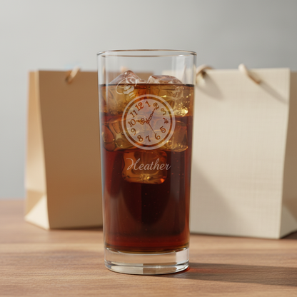 A clear highball glass filled with a dark drink, featuring a etched pocket watch design and the name 'Heather' on the front, with two gift bags in the background.