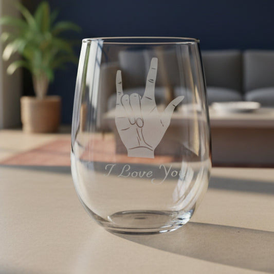 A clear stemless wine glass with an etched gray hand gesture and the words 'I Love You' on the front, shown standing on a wooden surface with a living room in the  background.