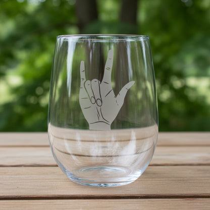 A clear stemless wine glass with a gray hand graphic on the front, resting on a wooden surface with trees in the  background.