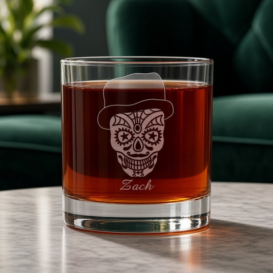 A clear rocks glass filled with amber liquid, featuring an etched sugar skull wearing a hat and the name 'Zach' near the bottom, set on a tabletop with a blurred green chair and plant in the background.