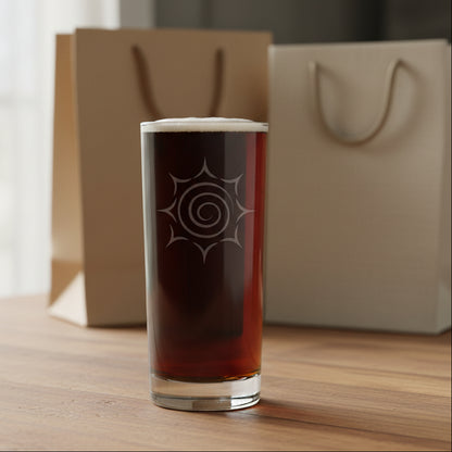 A tall, clear pint glass filled with a dark reddish-brown liquid, featuring an etched spiral sunburst design on the front, with a couple of gift bags in the background.