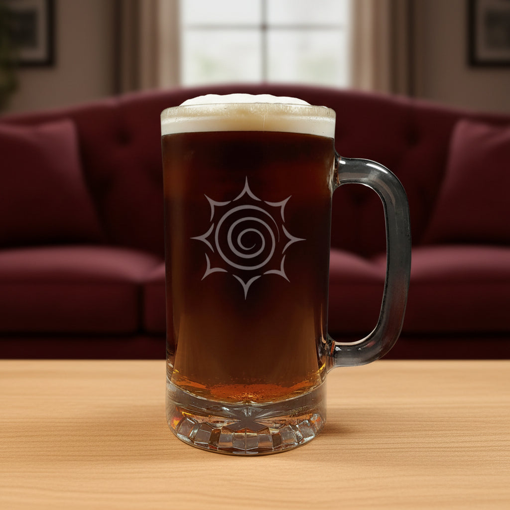 A clear glass beer mug with a handle, filled with dark amber beer and topped with white foam, featuring a etched spiral sunburst design on the front, set on a light wood surface against a red couch in the background.