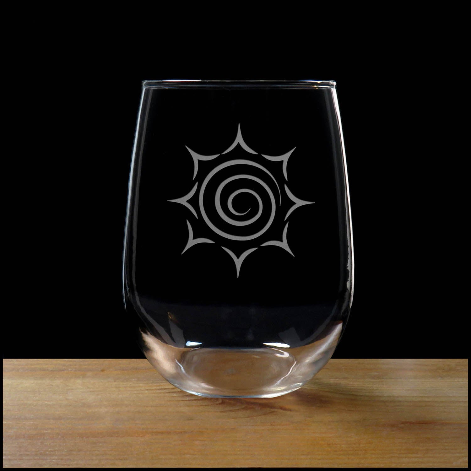 A clear stemless wine glass with a white etched spiral sunburst design, resting on a wooden surface against a solid black background.