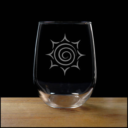A clear stemless wine glass with a white etched spiral sunburst design, resting on a wooden surface against a solid black background.