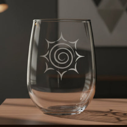 A clear stemless wine glass with a white etched spiral sunburst design, resting on a wooden surface with a living room in the background.