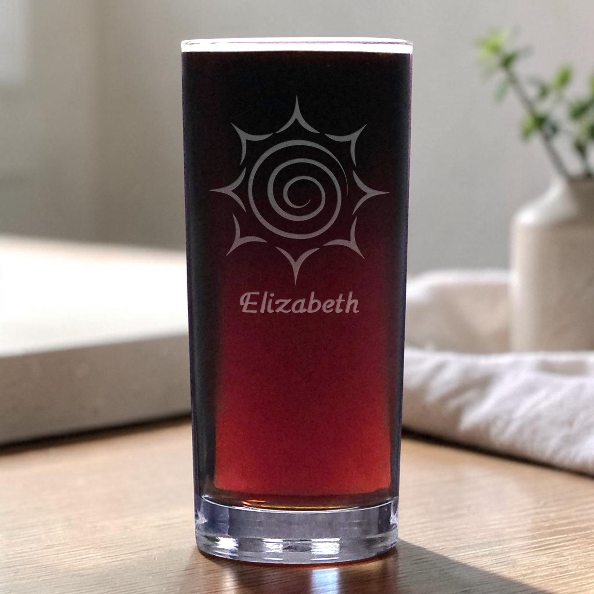 A clear glass pint glass filled with dark red wine, featuring a white etched sun design and the name 'Elizabeth' on the front, set on a wooden table in a softly blurred indoor setting.