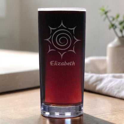A clear glass pint glass filled with dark red wine, featuring a white etched sun design and the name 'Elizabeth' on the front, set on a wooden table in a softly blurred indoor setting.
