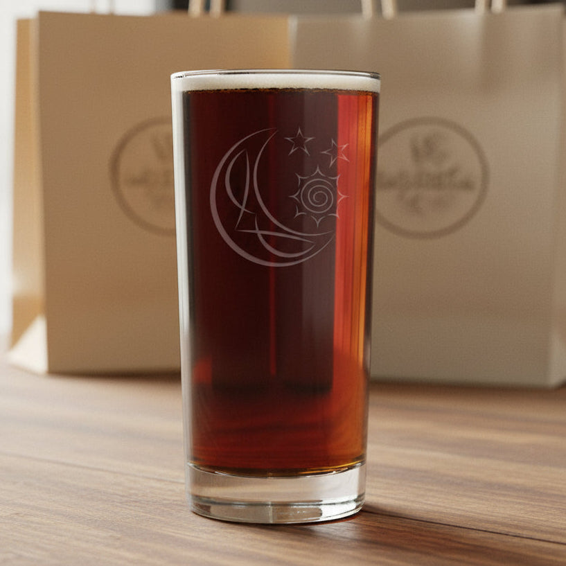A tall, clear cooler glass filled with a reddish-brown drink, featuring an etched crescent moon and sun design, and stars set against a  background of gift bags.