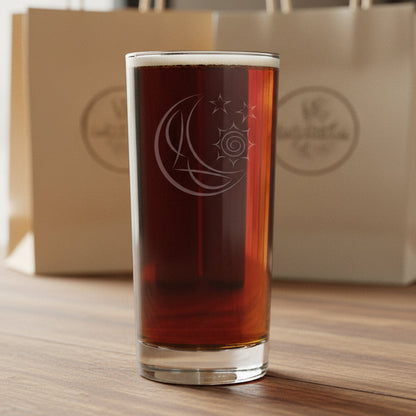A tall, clear cooler glass filled with a reddish-brown drink, featuring an etched crescent moon and sun design, and stars set against a  background of gift bags.