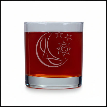 A clear glass rocks tumbler filled with an amber-brown liquid, featuring an etched crescent moon and star design on the front, set against a textured white background.