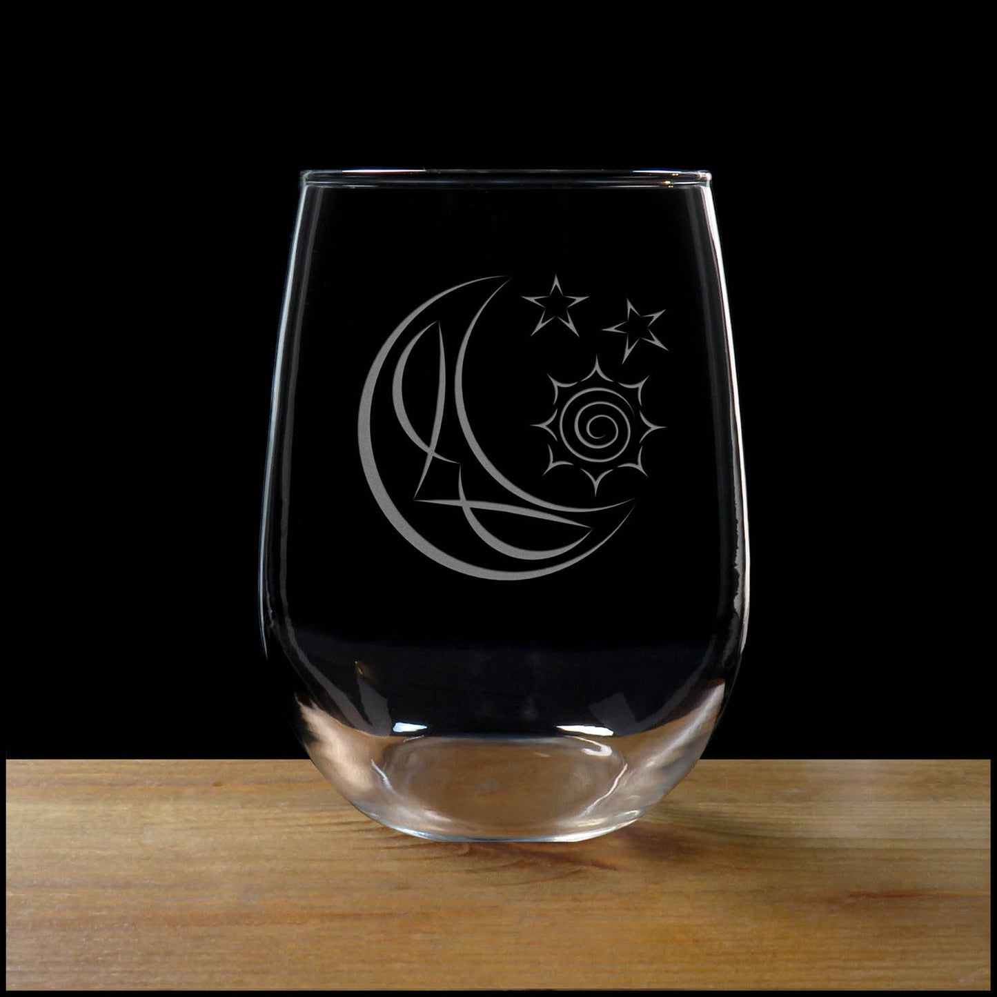 A clear stemless wine glass with an etched crescent moon and stars design, resting on a wooden surface against a solid black background.